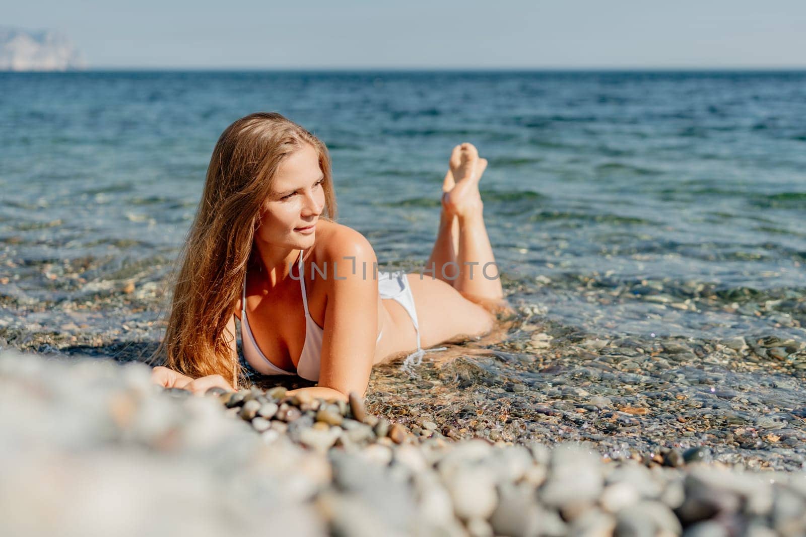 A woman is laying on the beach in a bikini. She is looking at the camera. The beach is rocky and the water is calm
