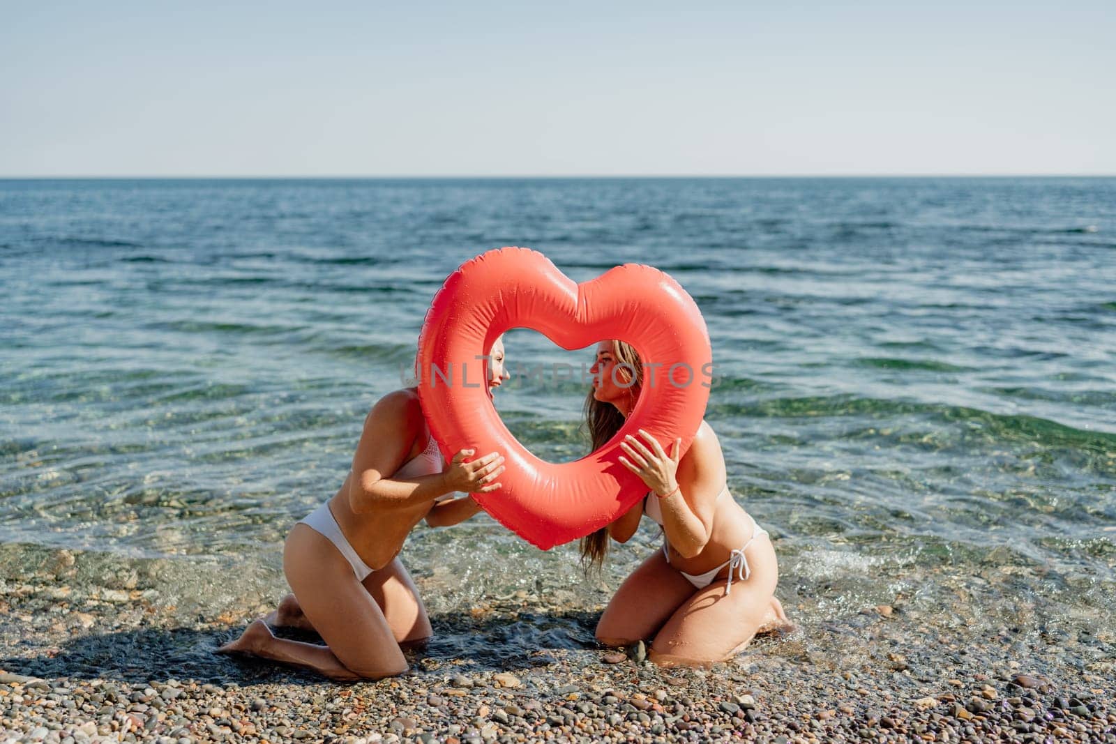 Two women are in the ocean, one holding a heart-shaped inflatable pool toy. Scene is playful and lighthearted, as the women are having fun and enjoying their time together