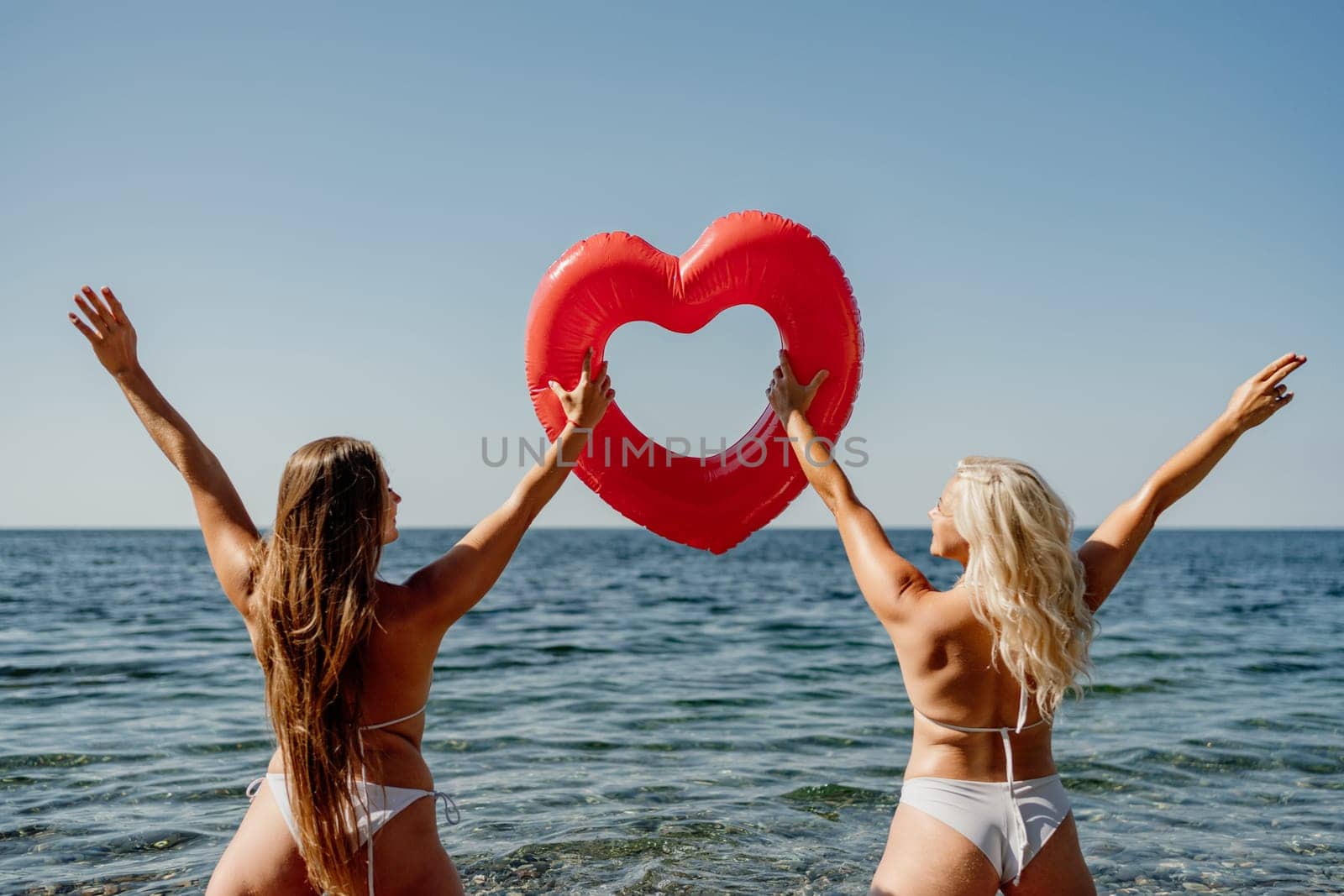 Two women are holding a red heart shaped inflatable raft in the ocean. They are smiling and seem to be enjoying themselves