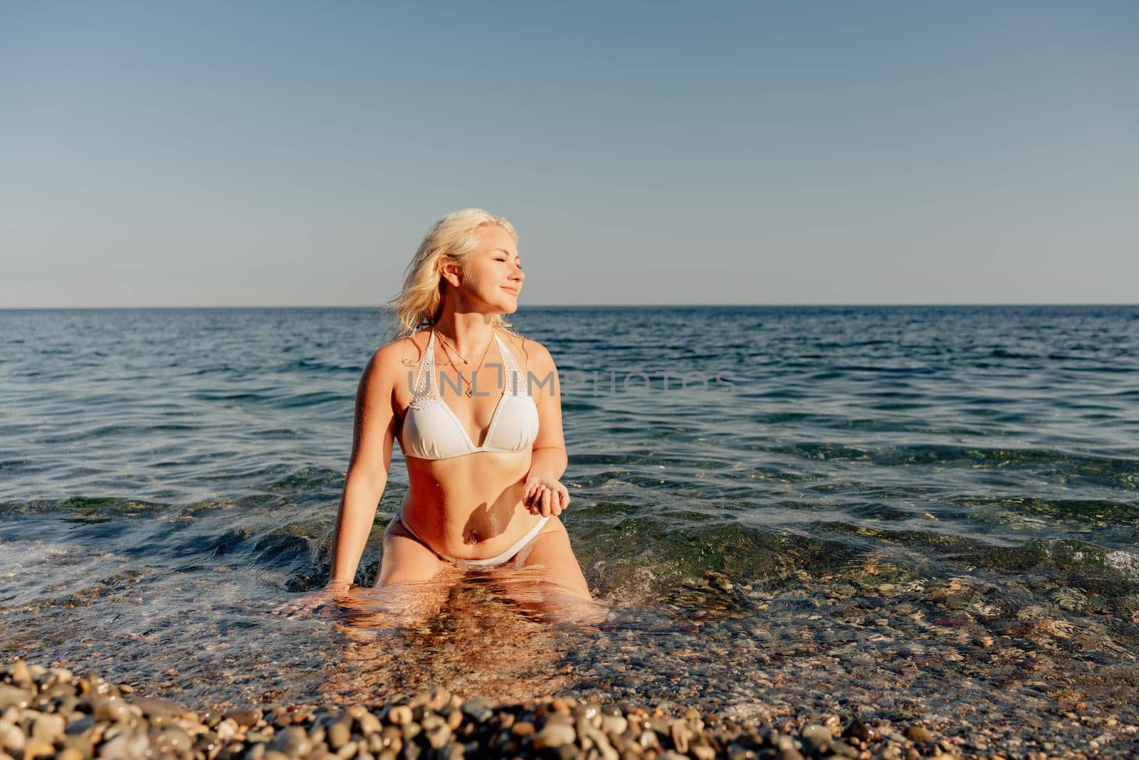 A woman in a bikini is sitting in the ocean. She is smiling and enjoying the water. Scene is happy and relaxed