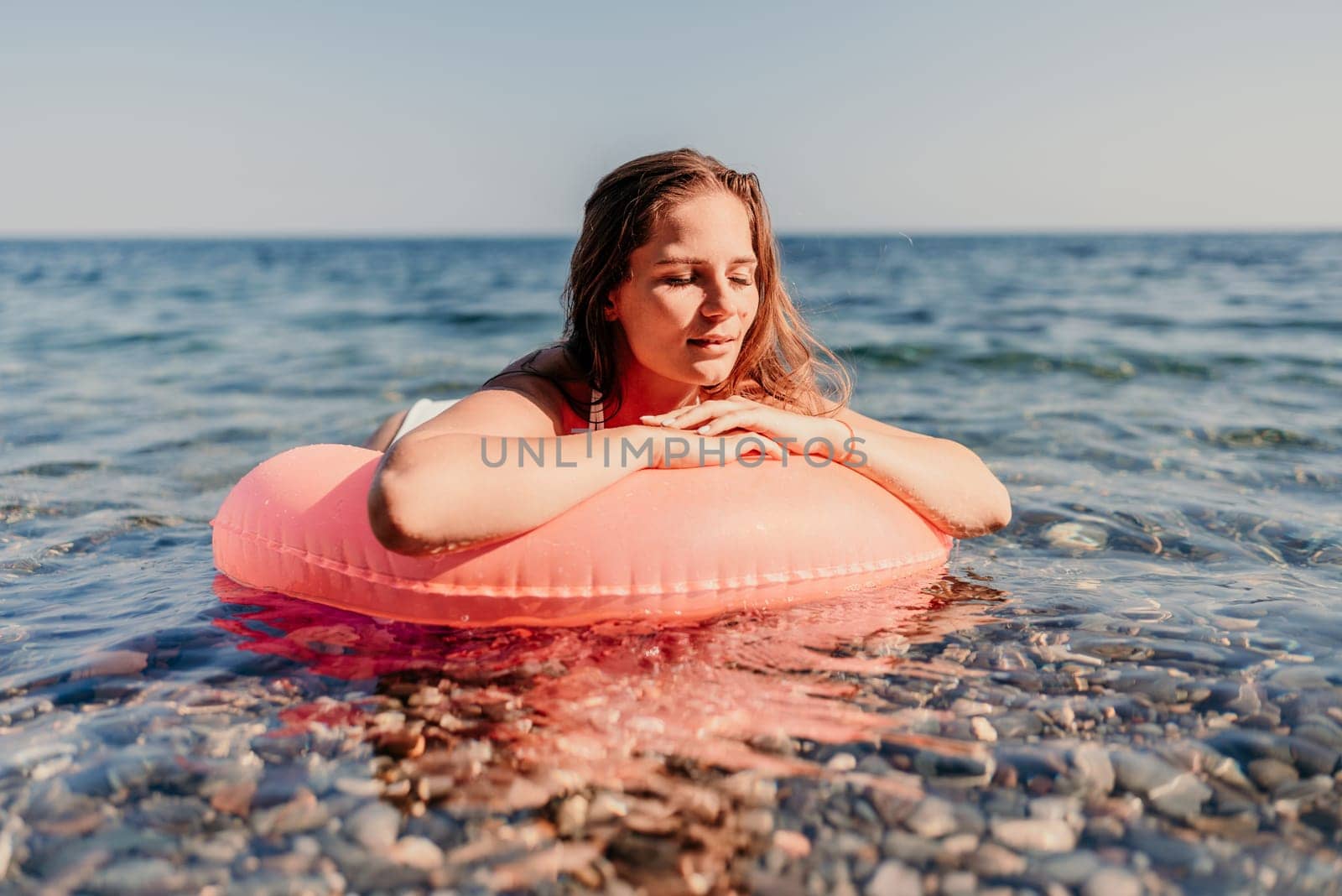 A woman is laying on a red inflatable ring in the ocean. The water is calm and the sky is clear. The woman is enjoying her time in the water