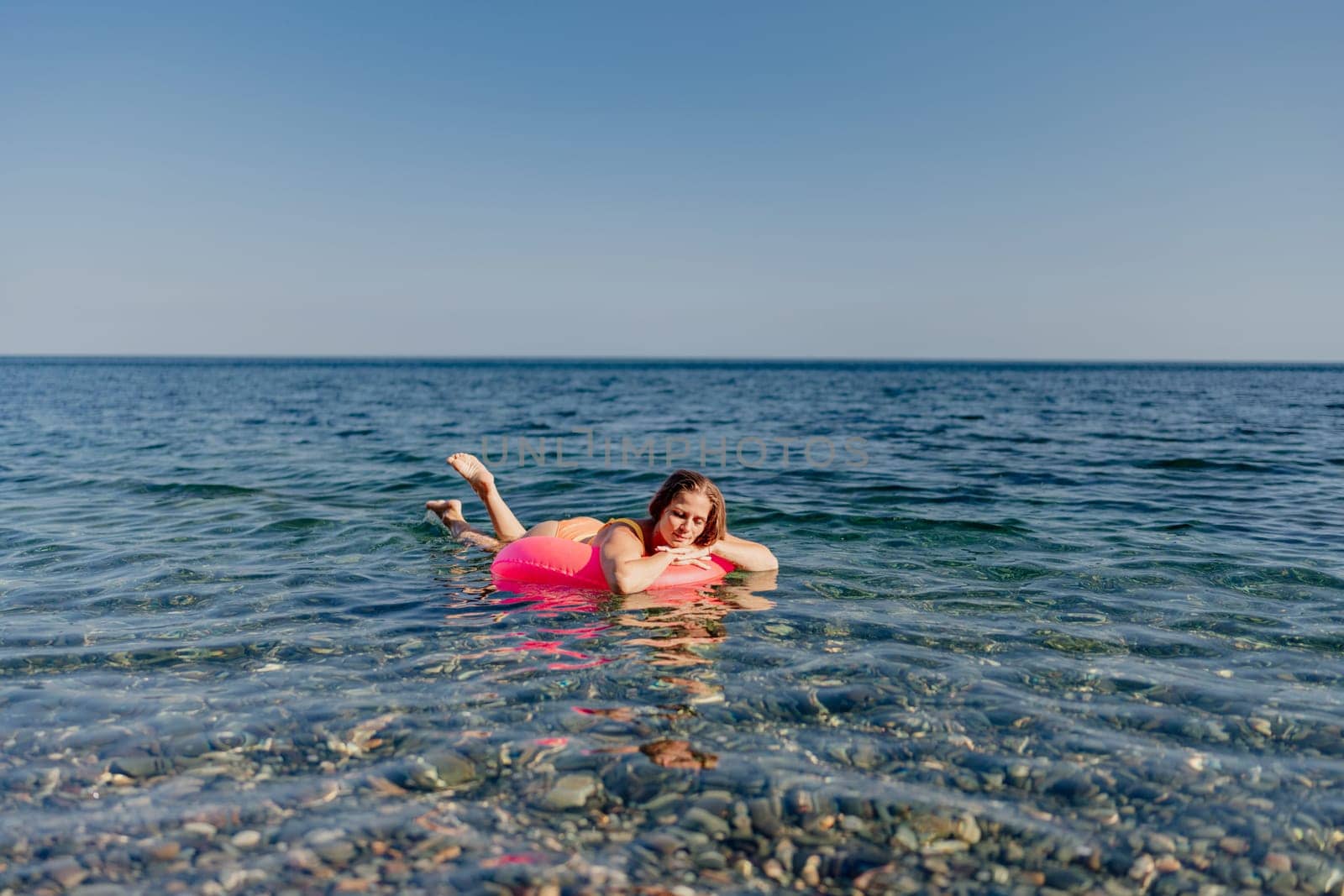 A woman is floating on a pink inflatable raft in the ocean. The water is calm and the sky is clear. The woman is enjoying her time in the water