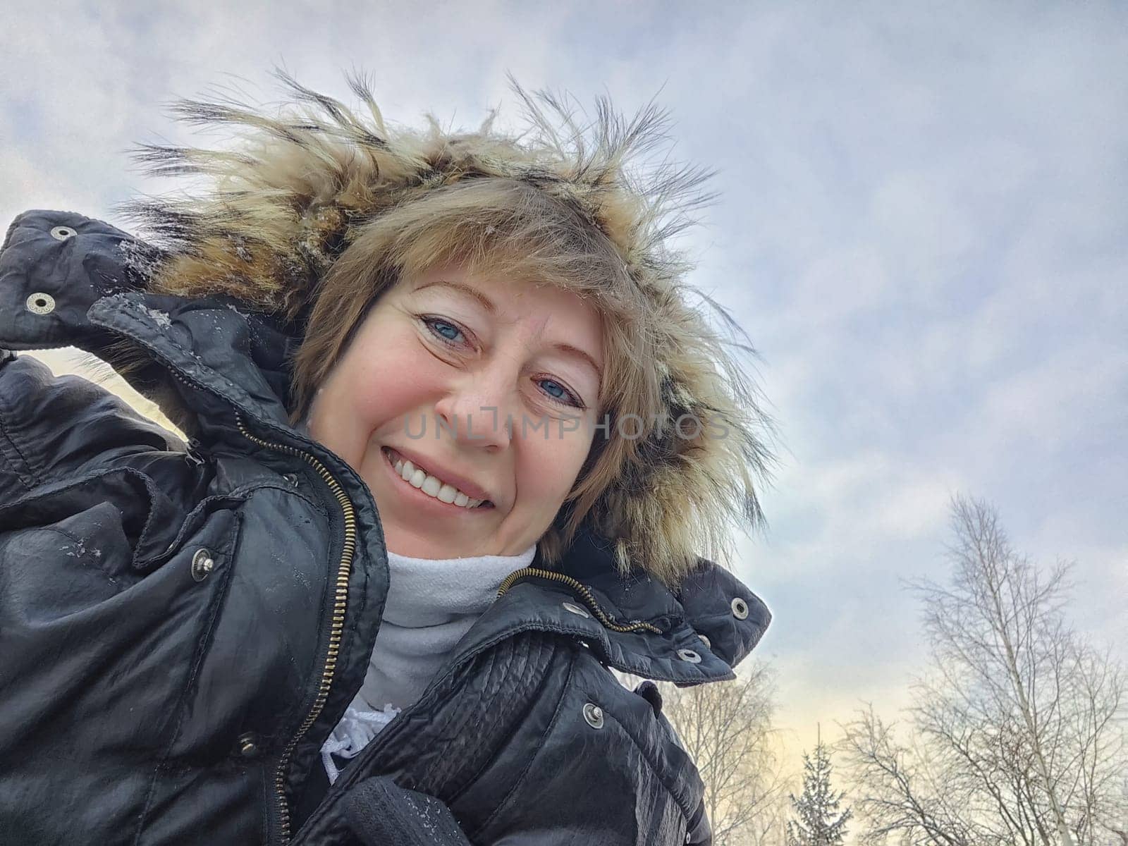 Happy, cheerful middle-aged woman in a warm jacket and a hood with fur taking selfie in winter outdoors. Female Traveler in Alaska, Lapland, Arctic, Siberia