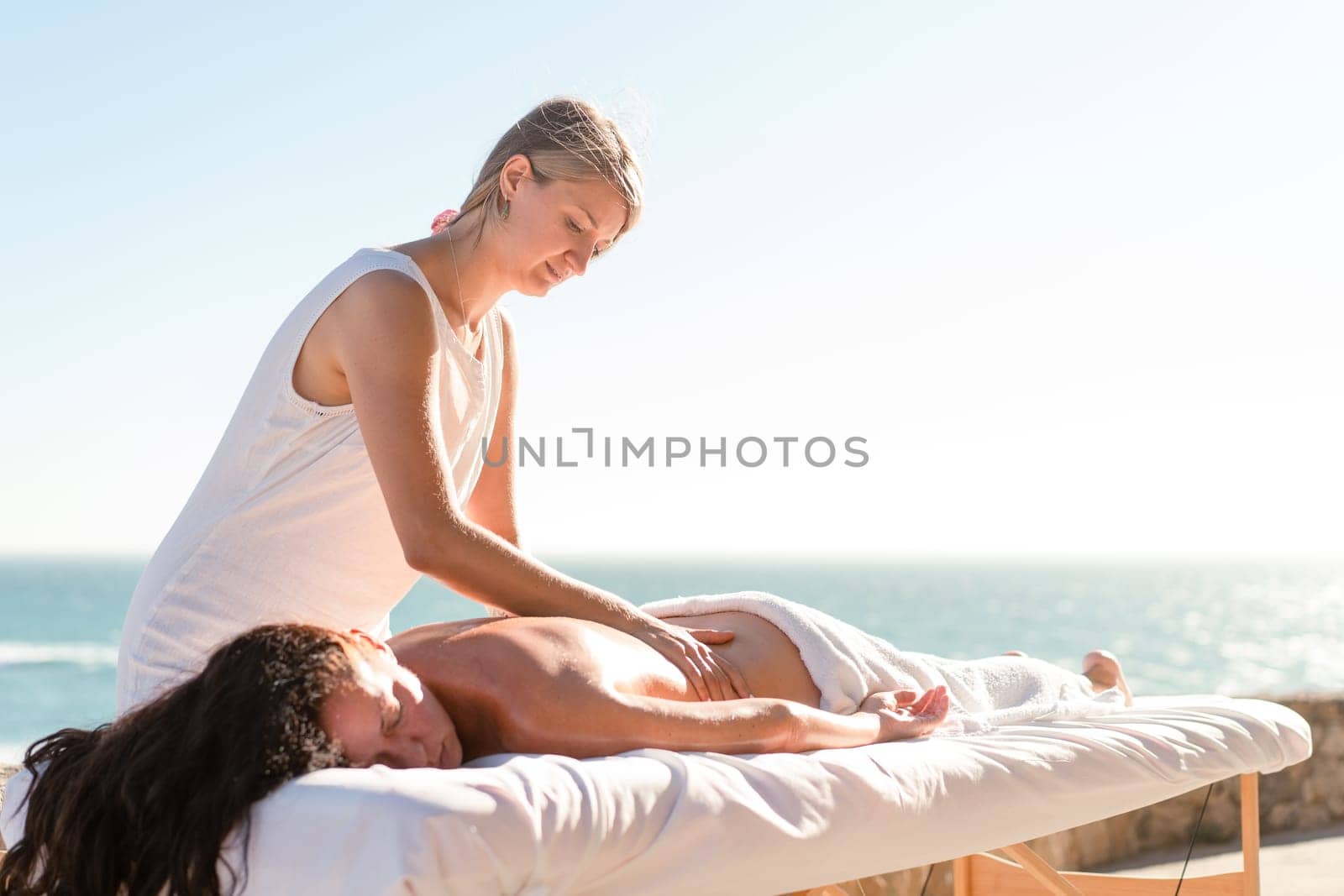 Female masseur massaging the back of relaxed female tourist on special table near sea. Brunette woman enjoying spa body treatment outdoors during summer vacation under clear sky.