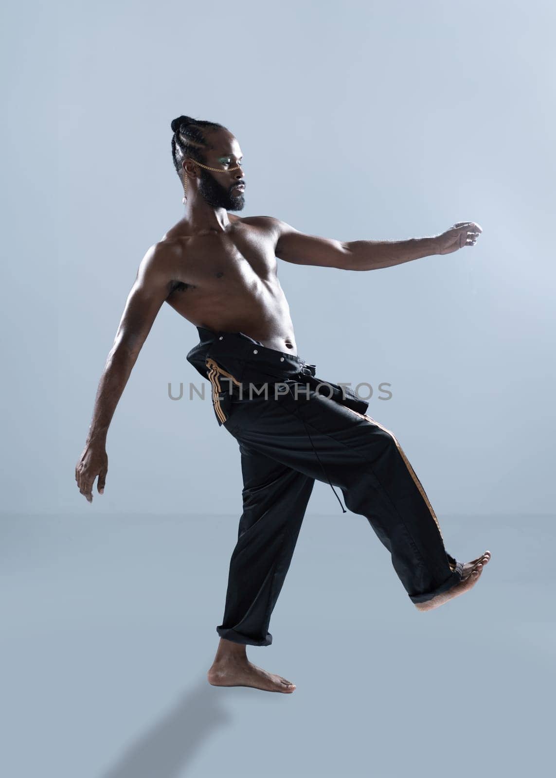 Side view of shirtless African American gay man confidently walking on light blue background. Full body shot showcases muscular LGBTQ man exuding confidence, wearing makeup and trendy pants.