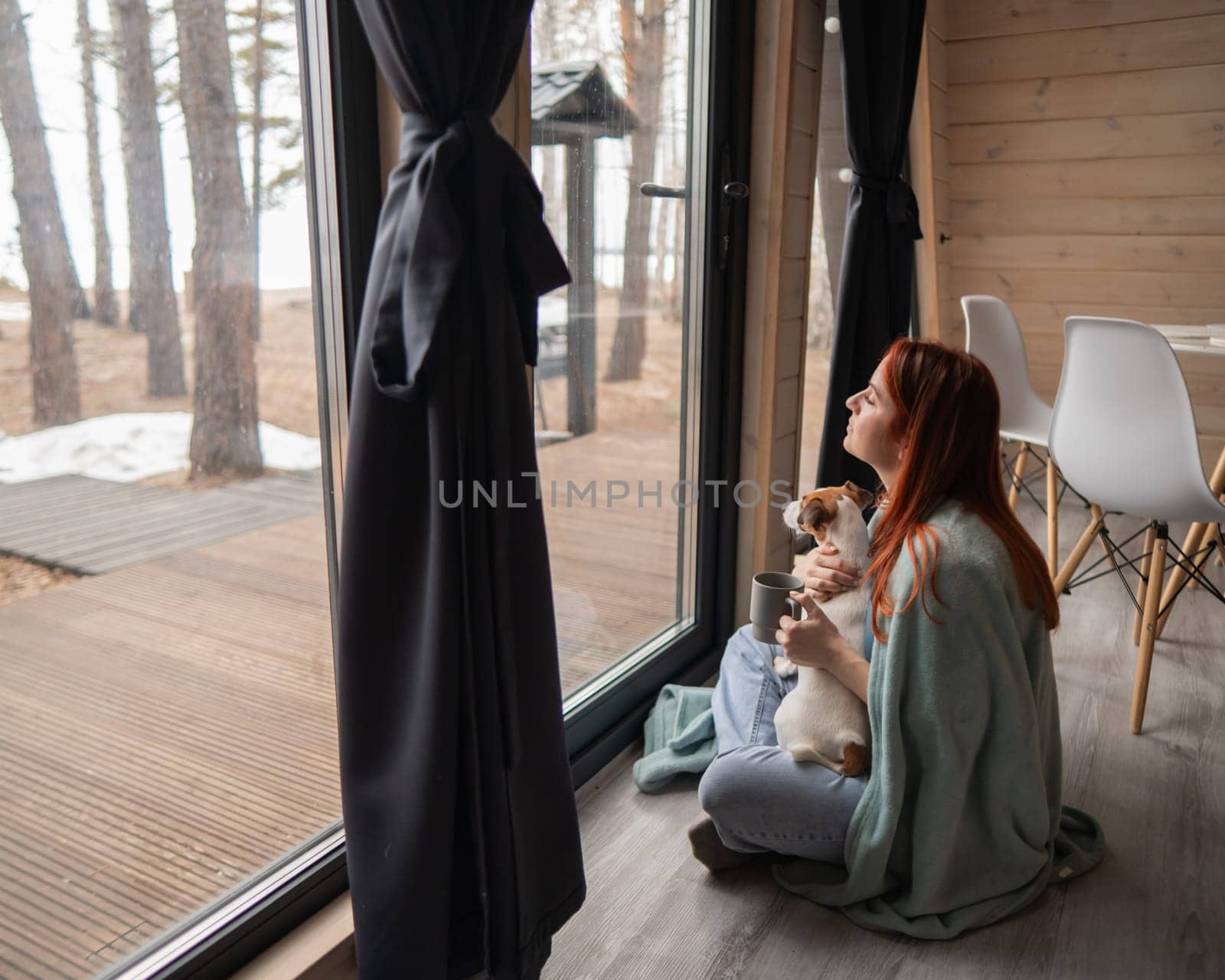 Caucasian woman wrapped in a blanket drinks coffee near the patio window hugging a Jack Russell terrier dog