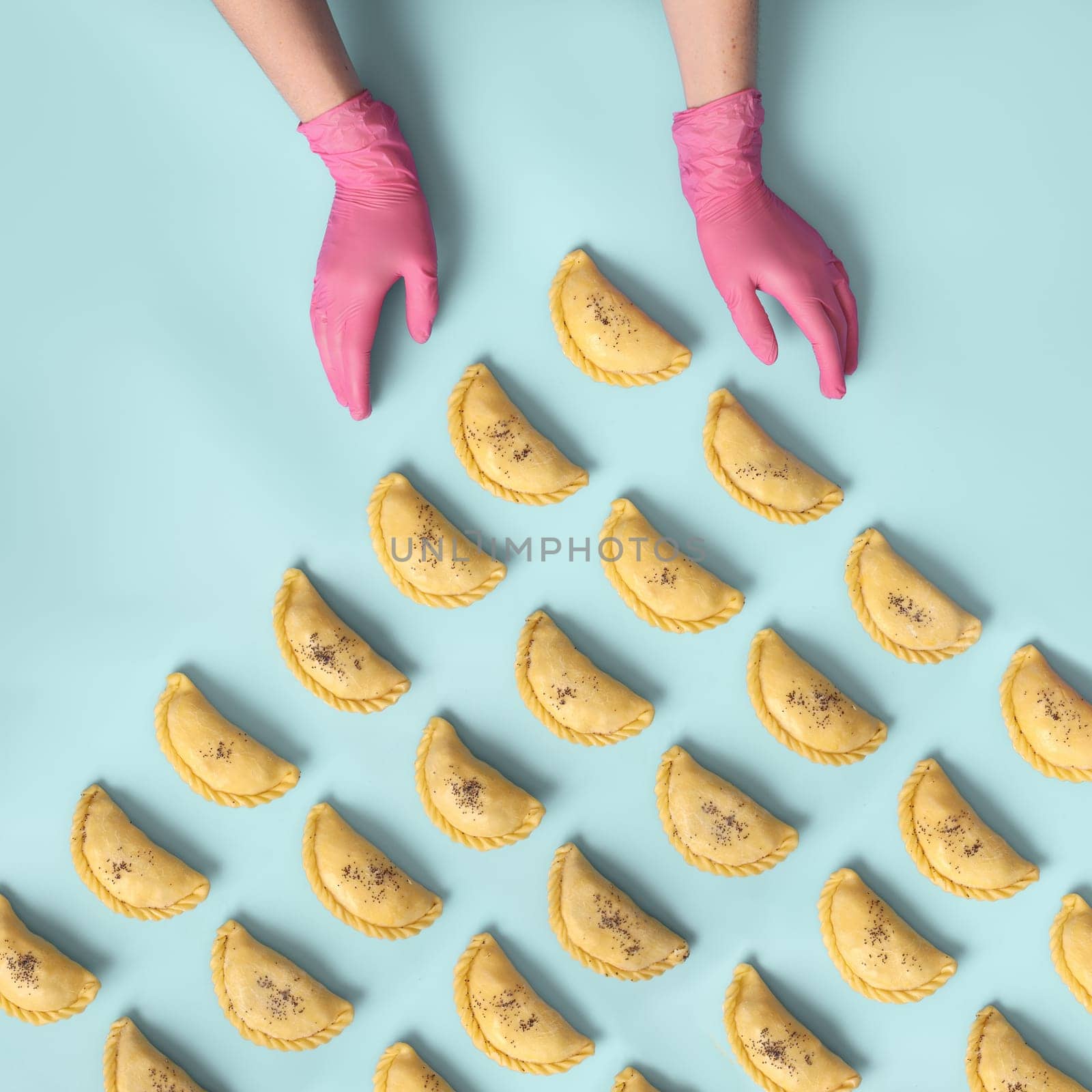 A baker's hands in a pink rubber gloves lays out raw samosa on blue background. Traditional Central Asian cuisine bakery, frozen semi-finished products.