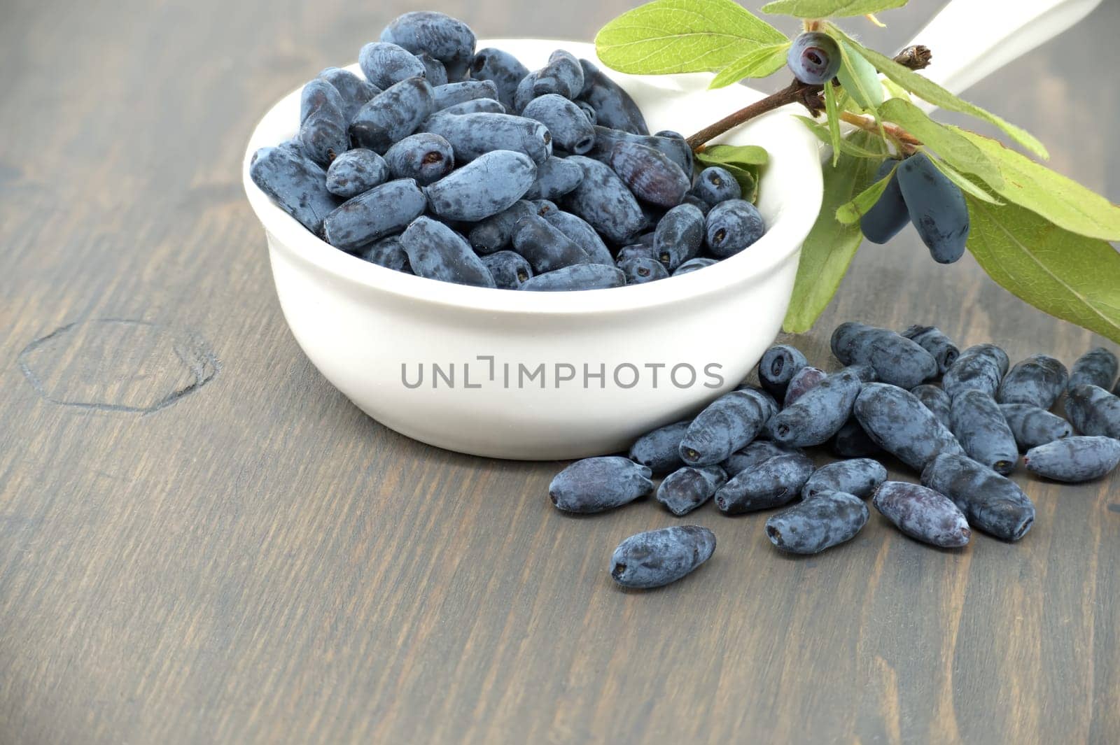 White porcelain bowl filled with blue honeysuckle berries is situated on a wooden table, some of berries spilling out onto the table near branch with fresh green leaves and berries
