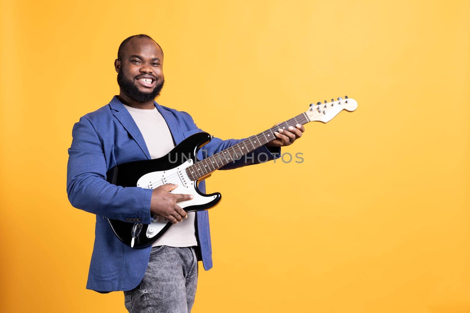 Portrait of happy man holding guitar, performing rock tunes during concert isolated over yellow studio background. Joyous artist performing compositions on electric musical instrument