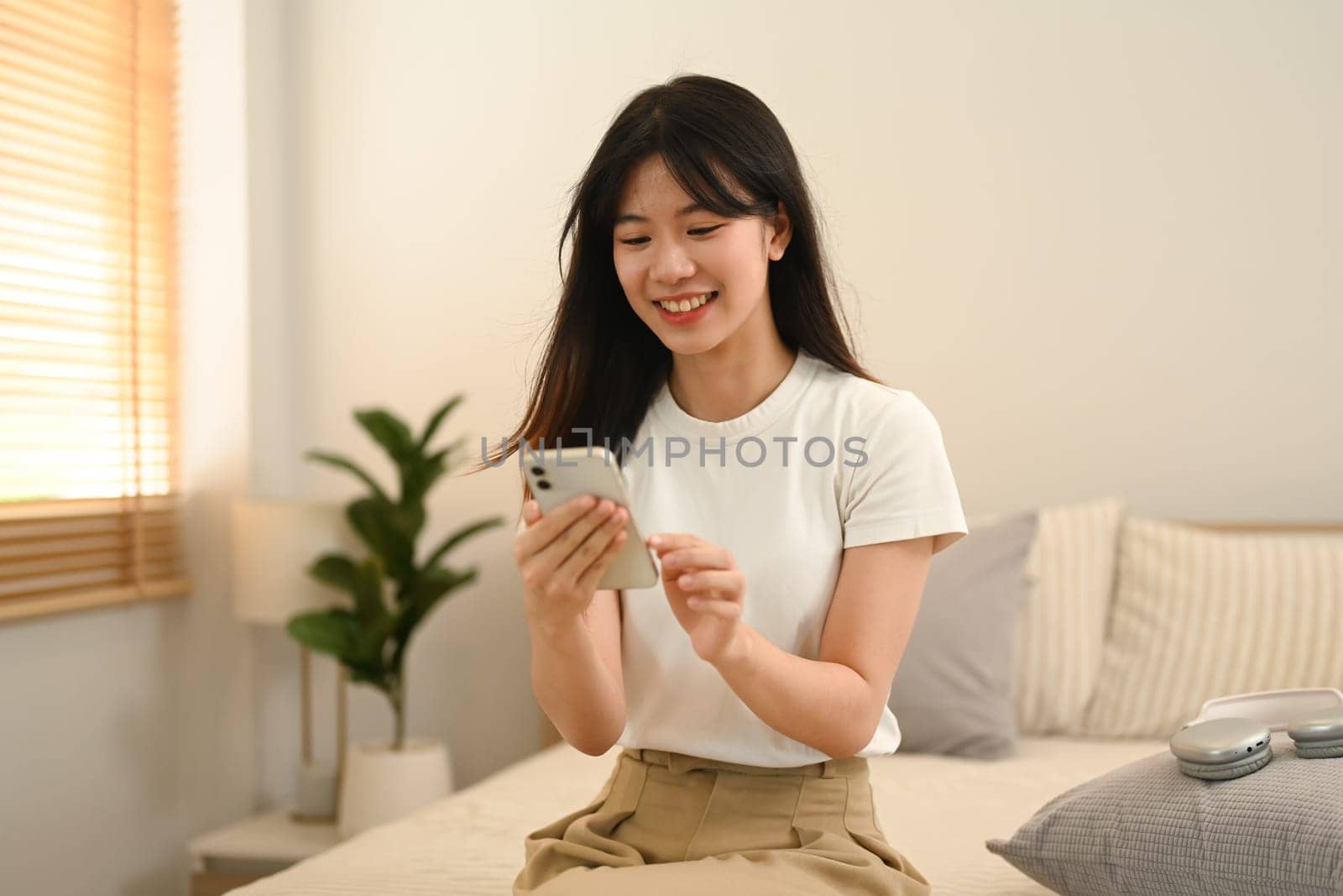 Pretty young woman using mobile phone while sitting on bed at home.