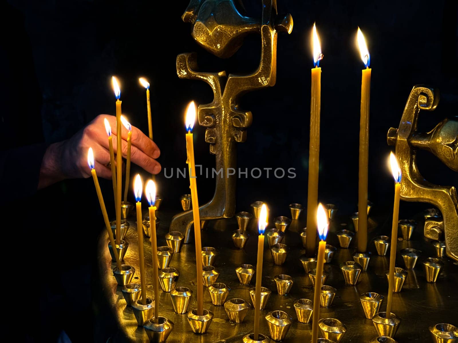 Hand lighting candles in dark room with golden candle holder. Religious or spiritual ceremony with candlelight. Lighting a candle in an Orthodox or Catholic church. High quality photo