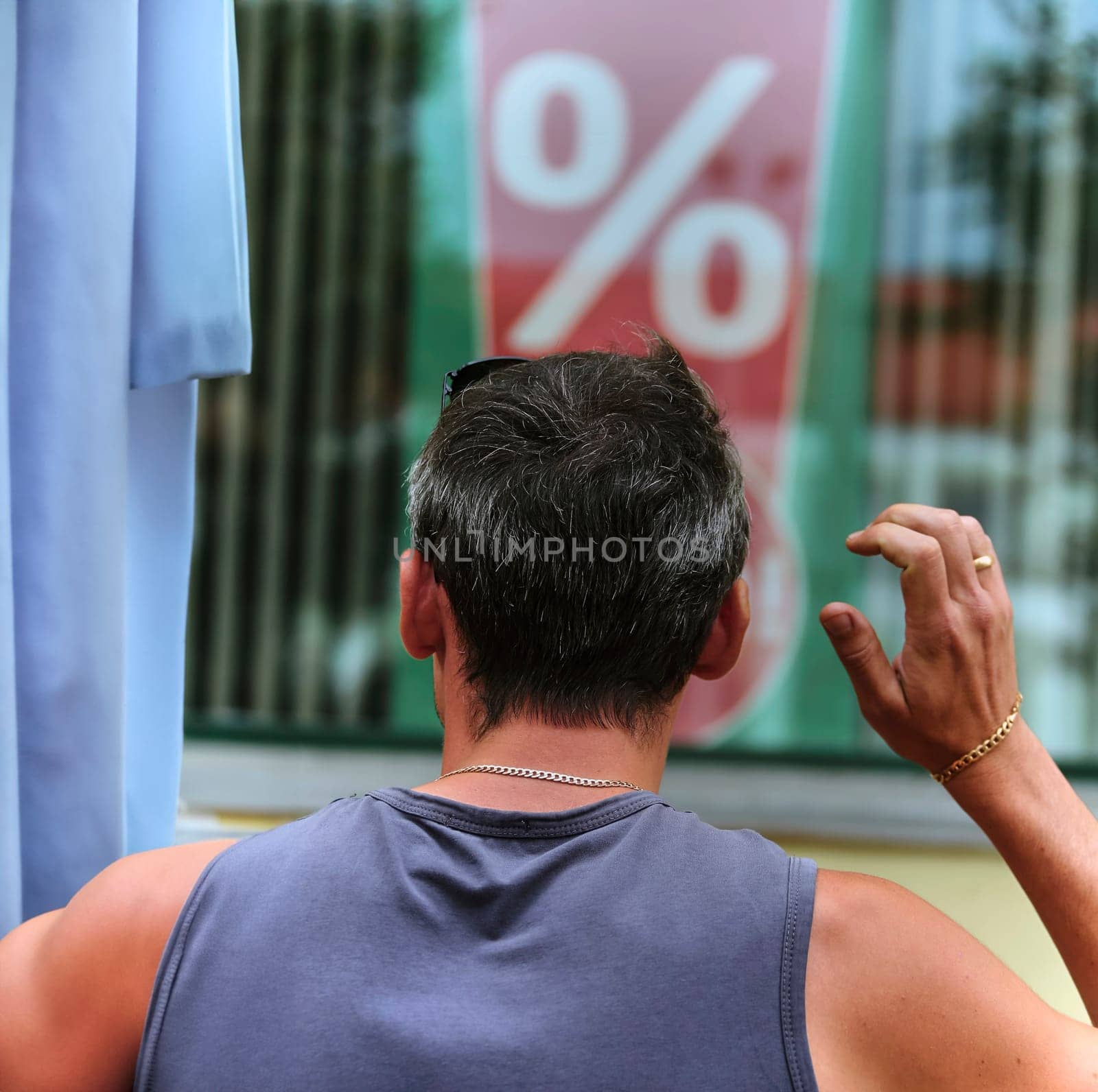 A man is standing outside a store, scratching his head as he observes a large percentage sign displayed in the window. His posture suggests he might be puzzled or considering the implications of the advertised discount, sale, or promotion.