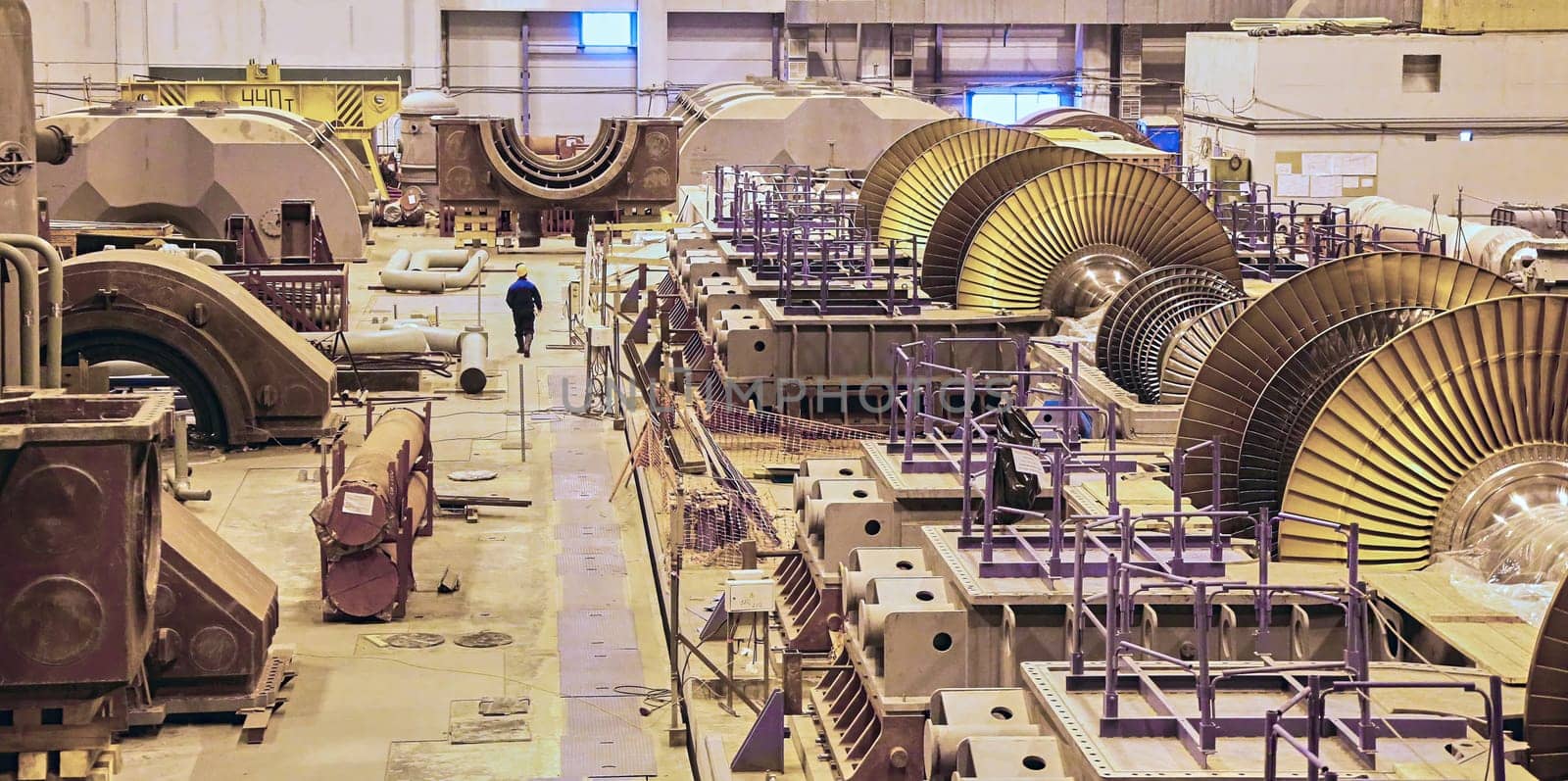 A lone worker walks through a vast factory floor, dwarfed by the imposing presence of massive turbine blades in various stages of assembly. The scene captures the raw power and intricate engineering involved in constructing these essential components of modern energy infrastructure.