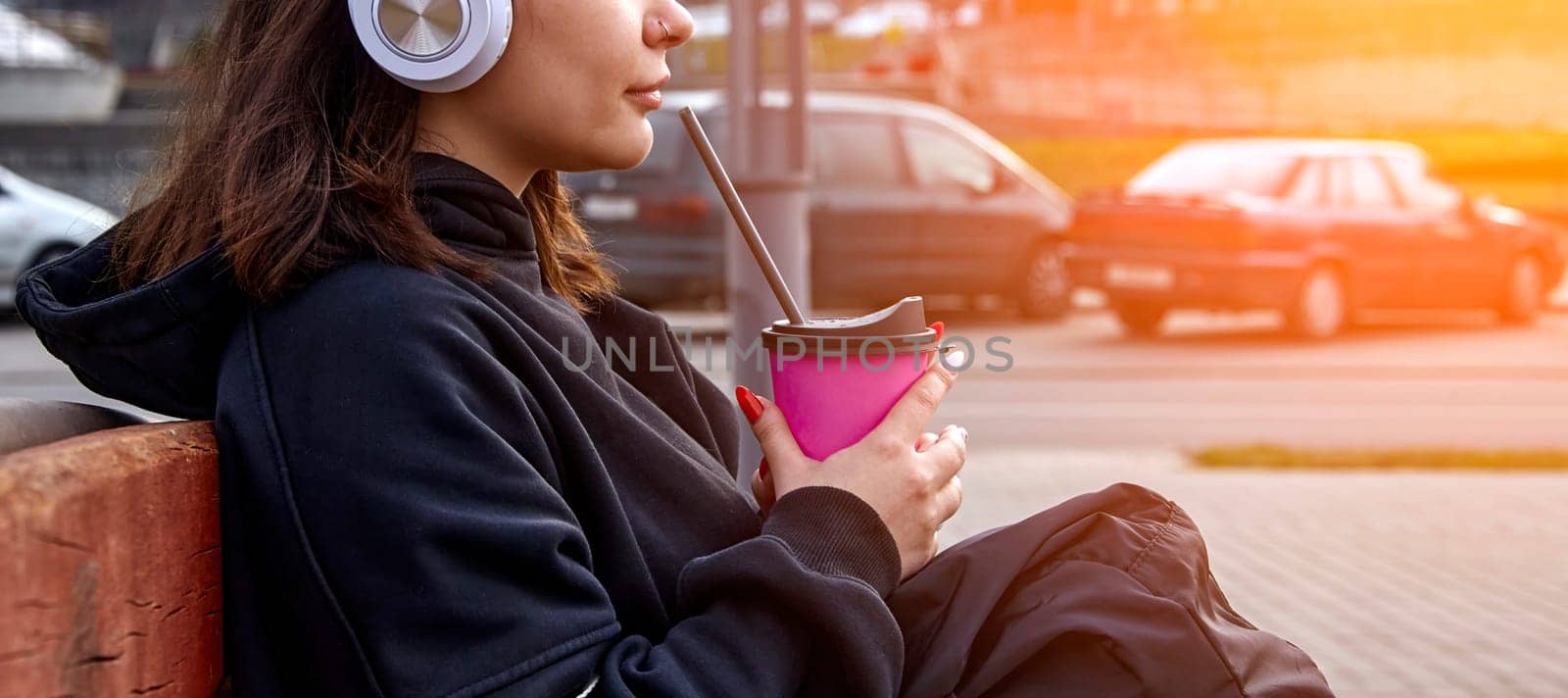 A young woman sits on a bench in an urban setting, enjoying a refreshing beverage on a warm afternoon. She wears headphones and seems engrossed in her own thoughts as she takes a break amidst the citys bustling energy. The sun shines brightly in the background, creating a warm and inviting atmosphere.