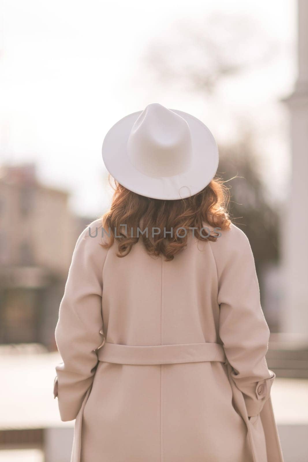 Back view of happy woman wearing hat and coat walking down street on sunny spring day. People, lifestyle, travel and vacations concept.