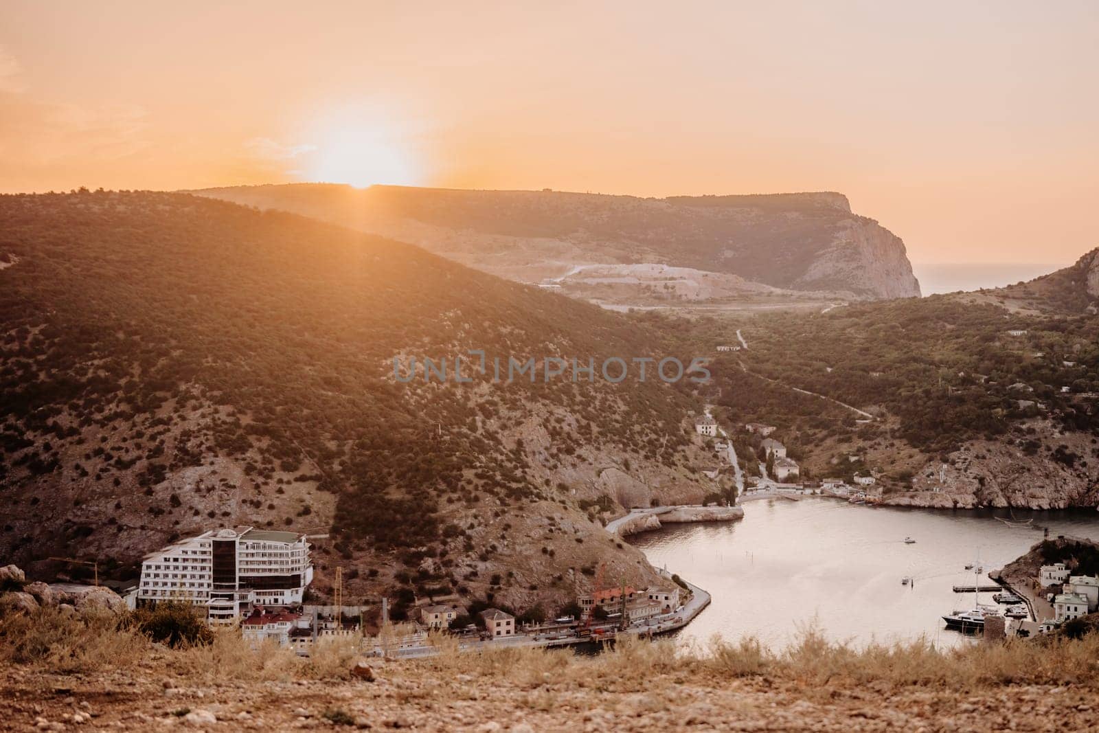 A beautiful sunset over a mountain with a small town in the distance. The sun is setting behind the mountains, casting a warm glow over the landscape. The water in the valley is calm and peaceful