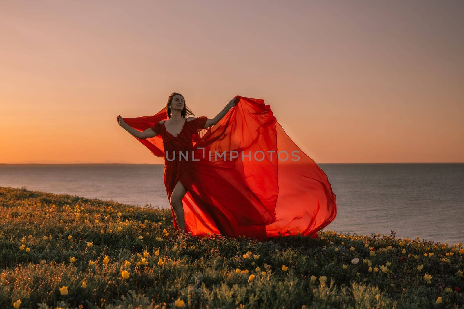 woman red dress is standing on a grassy hill overlooking the ocean. The sky is a beautiful mix of orange and pink hues, creating a serene and romantic atmosphere