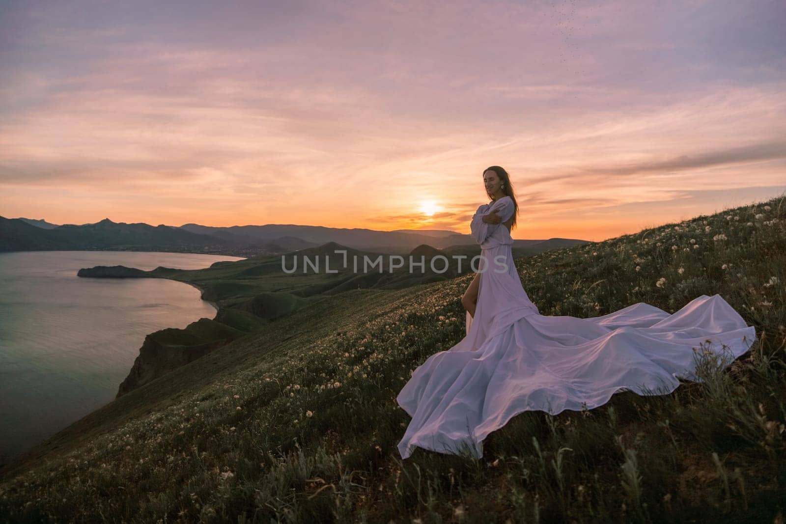 A woman in a long white dress stands on a hill overlooking a lake. The sky is a mix of orange and pink hues, creating a serene and romantic atmosphere. The woman's dress billows in the wind