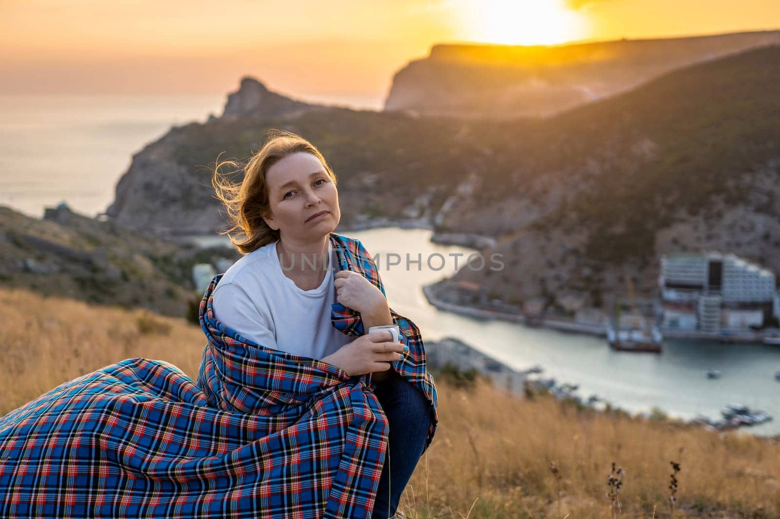 A woman is sitting on a hill with a blanket wrapped around her. She is looking out at the ocean