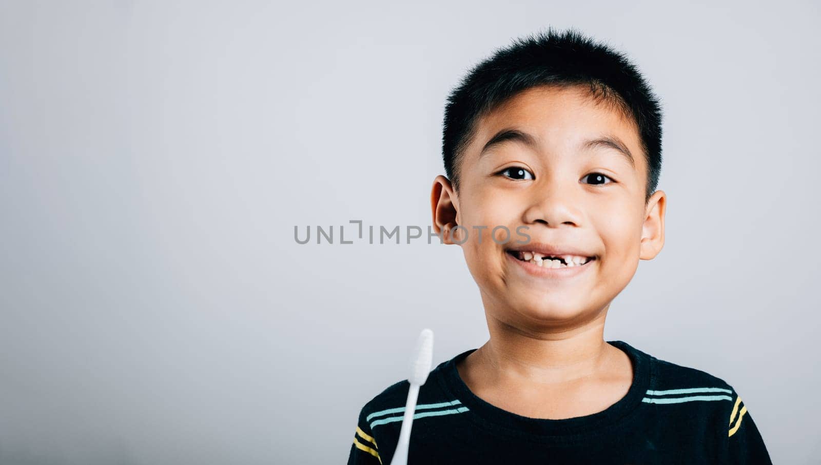 Isolated on a white background a cute Asian kid smiles holding a toothbrush emphasizing dental hygiene and the joy of learning without an upper milk tooth. Children dentist routine