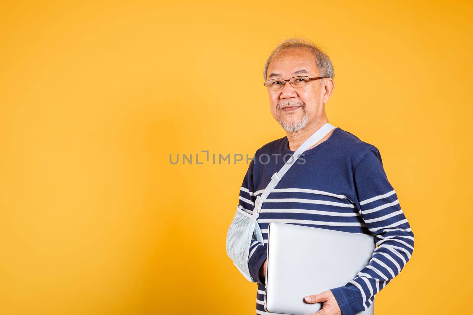 Portrait Asian old business man cast his arm hold laptop computer studio isolated yellow background, elderly man accident broken arm wear splint treatment while working by Sorapop