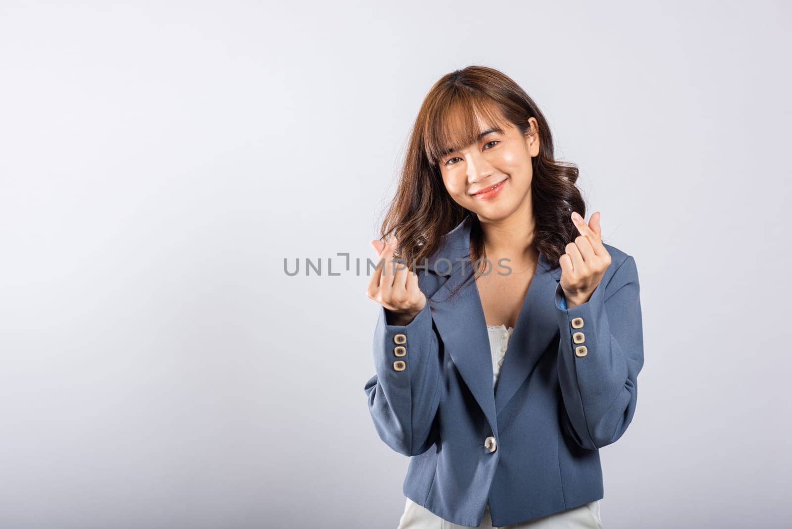 Confident business woman stands with a smile, creating a mini heart sign with her finger on a white background. Asian portrait of a beautiful young female sending love for Valentine's Day, studio shot