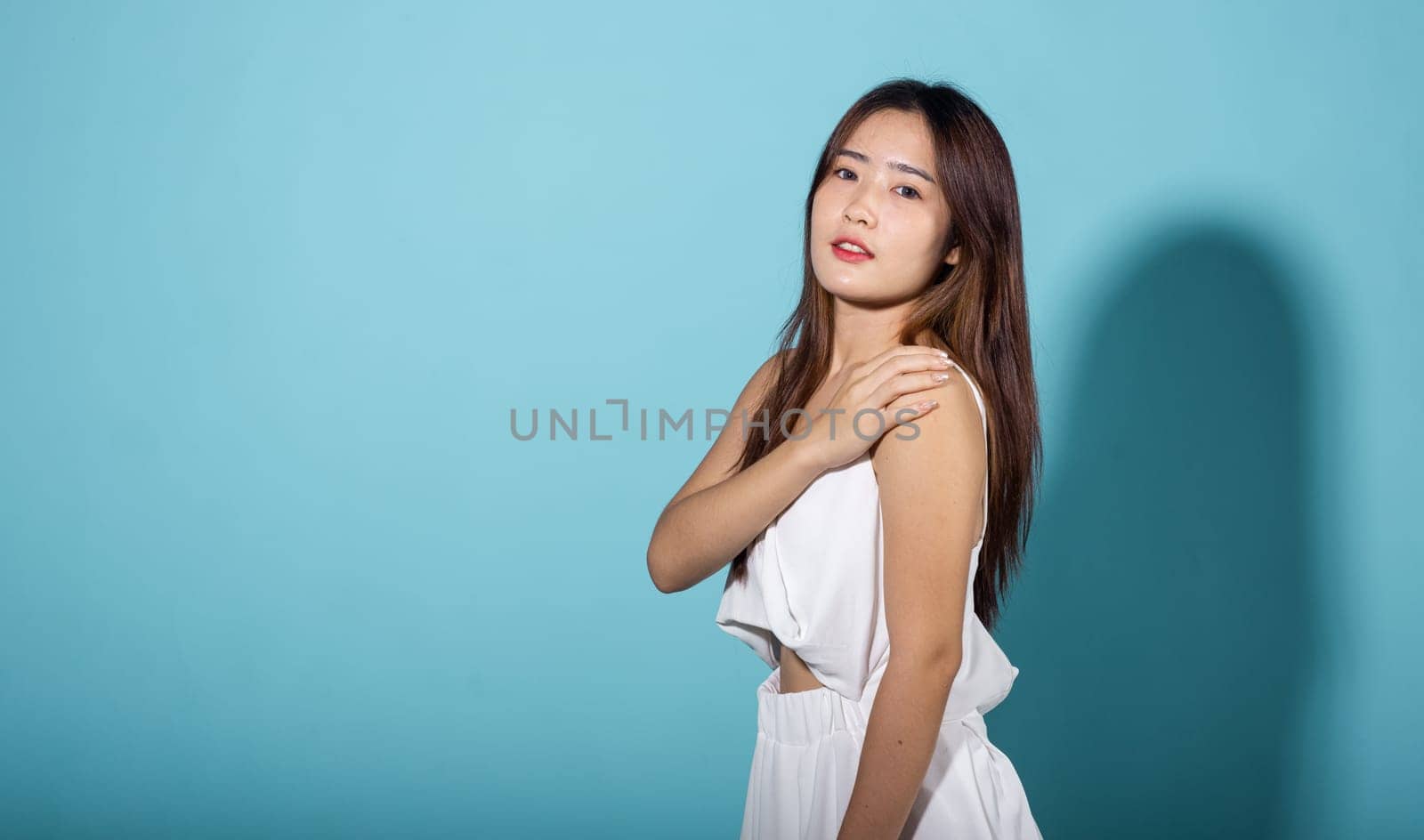 Female raising arm and hand in protection and block sunshine in studio shot isolated on blue background, Asian young woman smile hand up cover face protect sun light and UV, sunscreen cream advertise