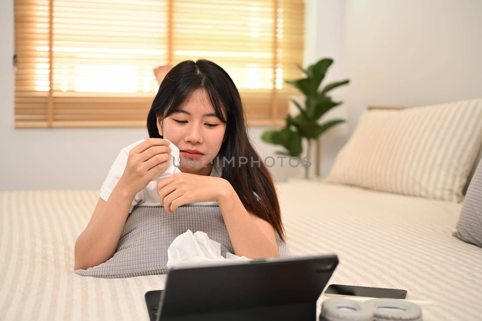 Young Asian woman crying and wiping tears while lying on bed watching dramatic movie.