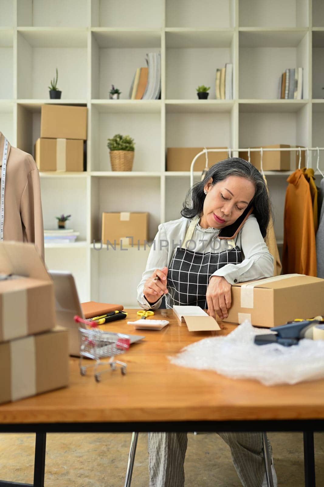 Senior female small business owner having phone conversation and calculating expense at desk.