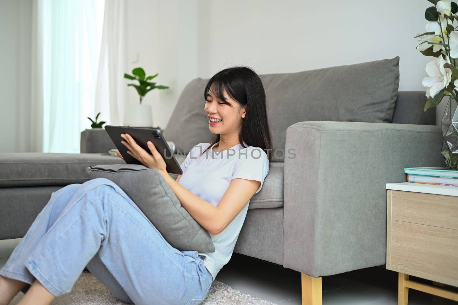 Happy young woman in casual clothing sitting on floor at home and using digital tablet.