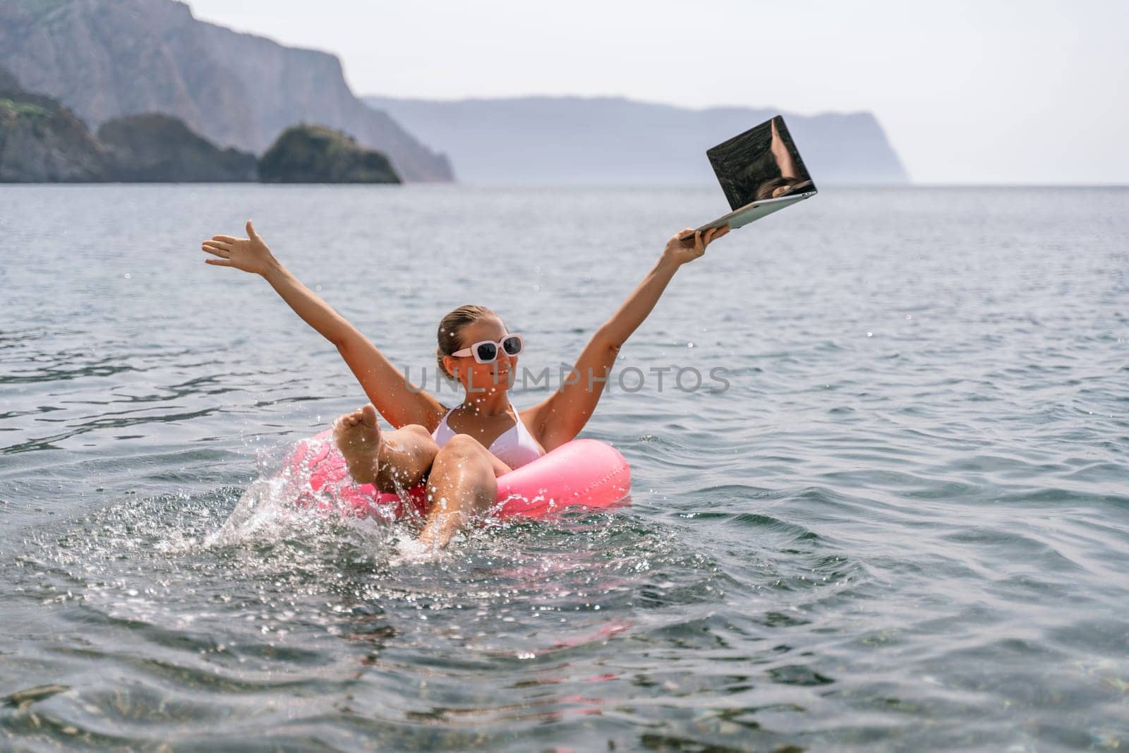 A woman is floating in a pink inflatable raft with a laptop in her hand. She is smiling and she is enjoying her time in the water