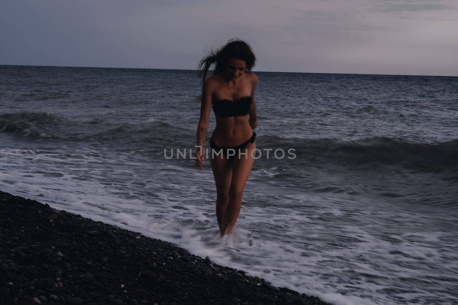happy young girl in a swimsuit runs on sea waves on beach on a summer evening. Photo with a blur from a long exposure to give an authentic atmosphere