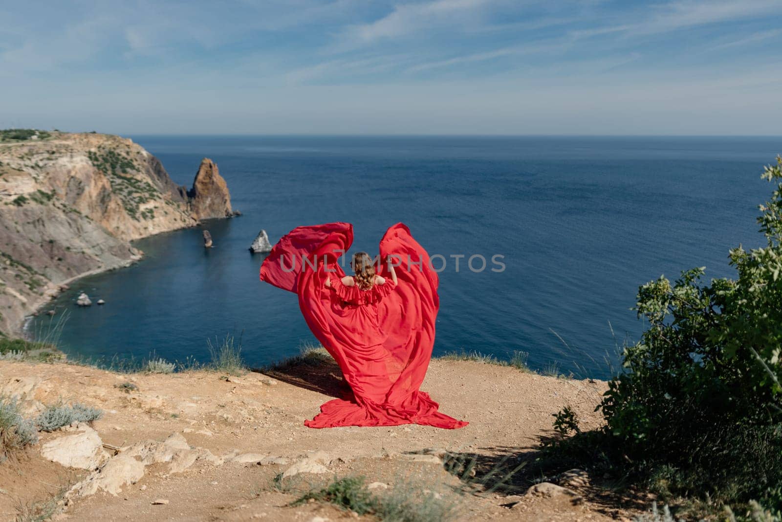 A woman in a red dress is standing on a rocky beach, with the ocean in the background. She is wearing a red dress and her hair is flowing in the wind. The scene is serene and peaceful