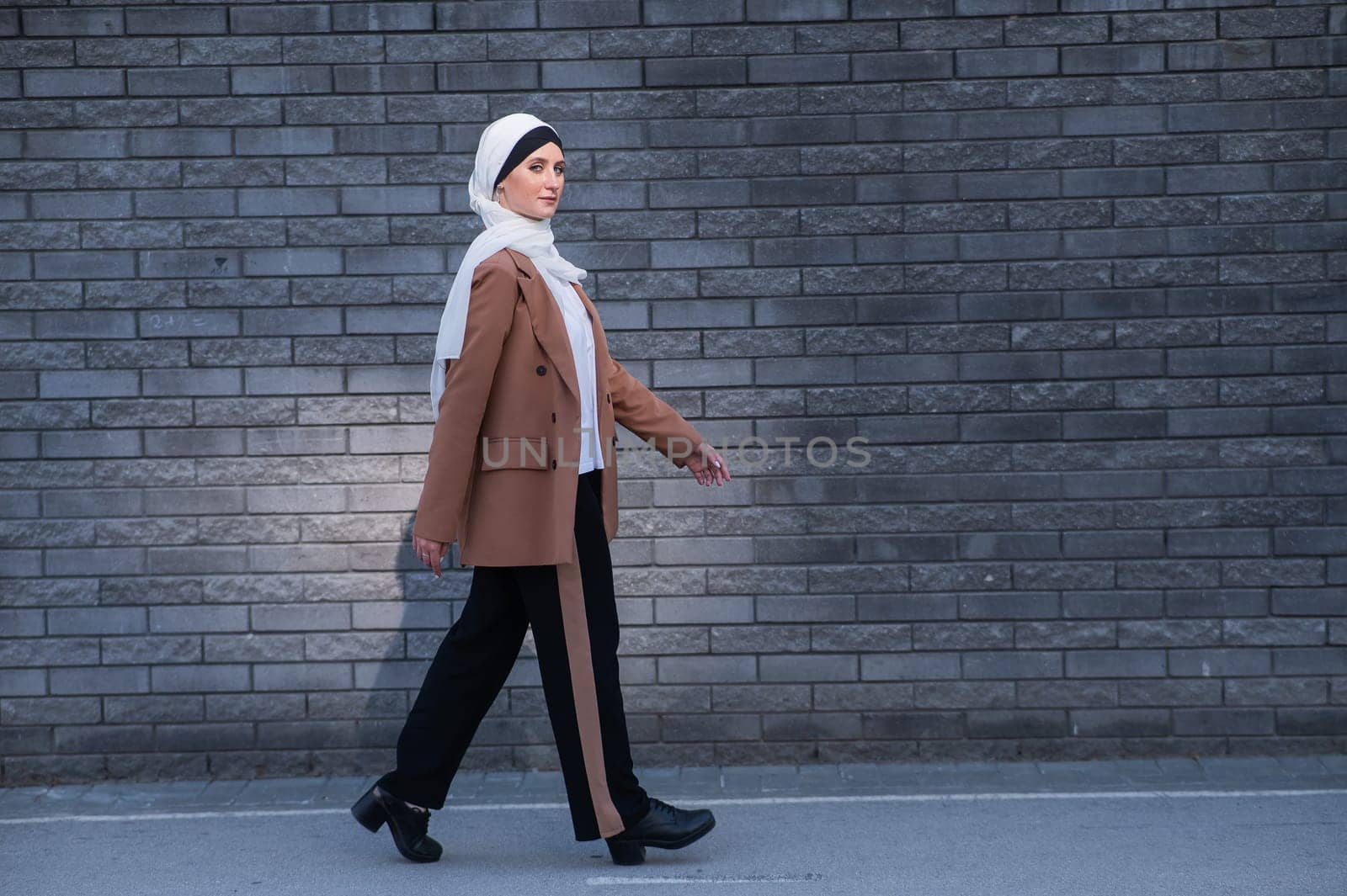 A young woman dressed in a hijab and a business suit walks along a brick wall