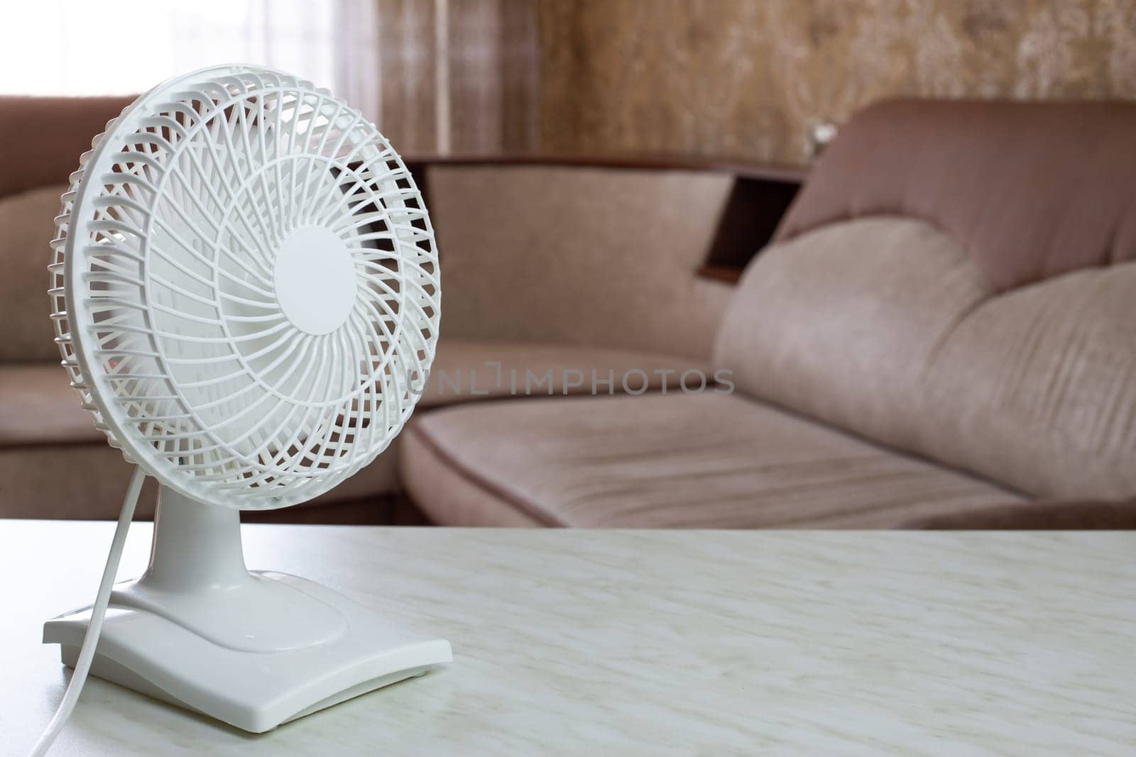 Tabletop compact white plastic fan with included blades disperses cold air throughout the home room against the backdrop of the sofa on hot day