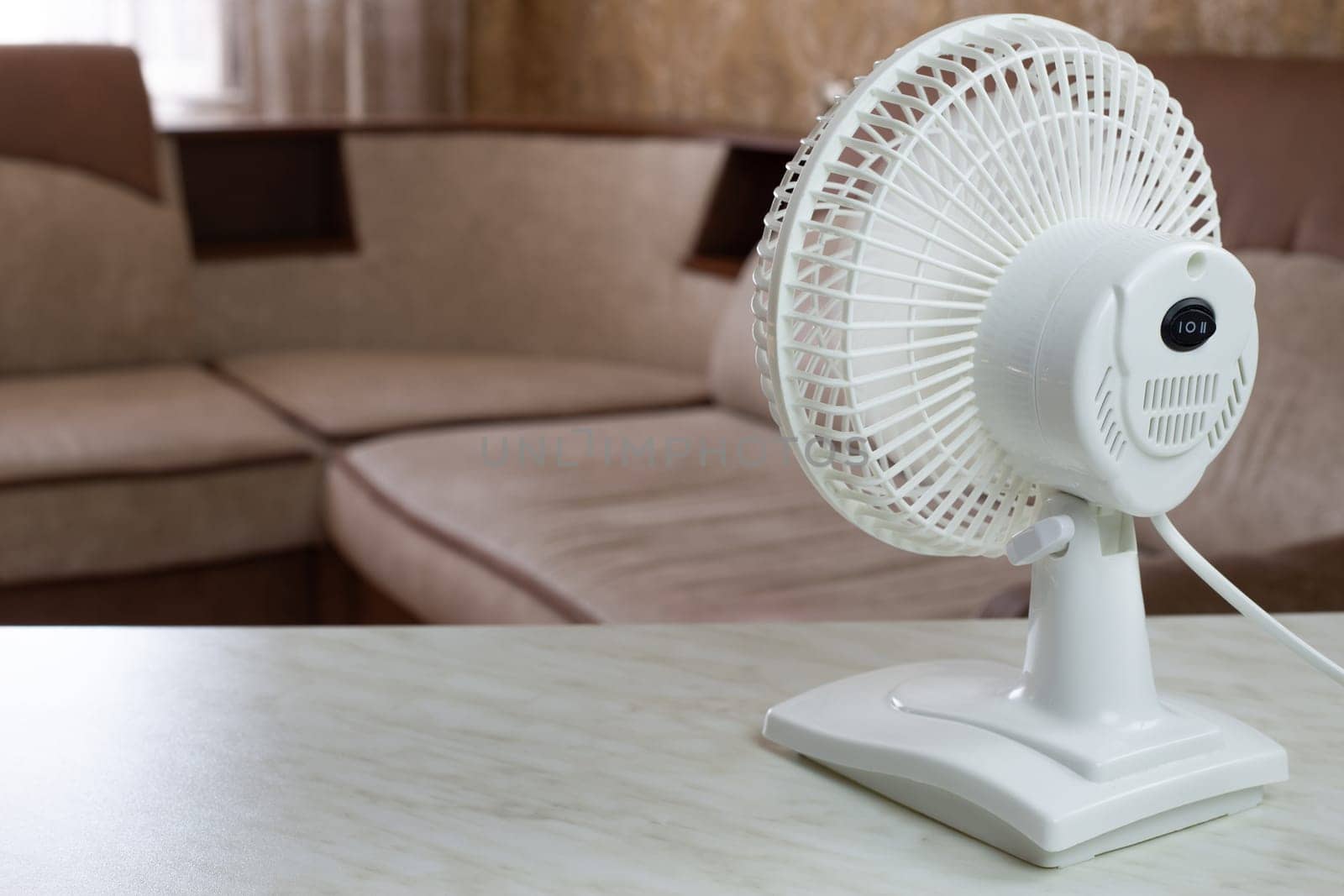 White fan on the table is turned on to cool the air in room rear view by timurmalazoniia