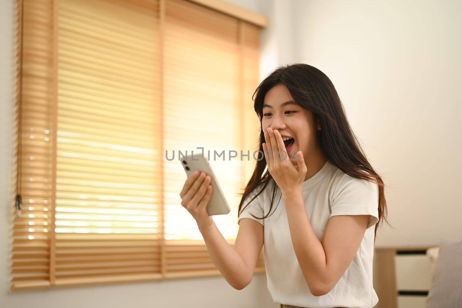 Shocked young asian lady looking at smartphone screen excited by unexpected notification.