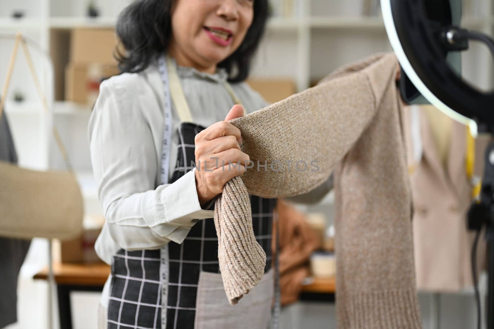 Senior female small business owner showing and review product in front of smartphone live streaming at her shop.