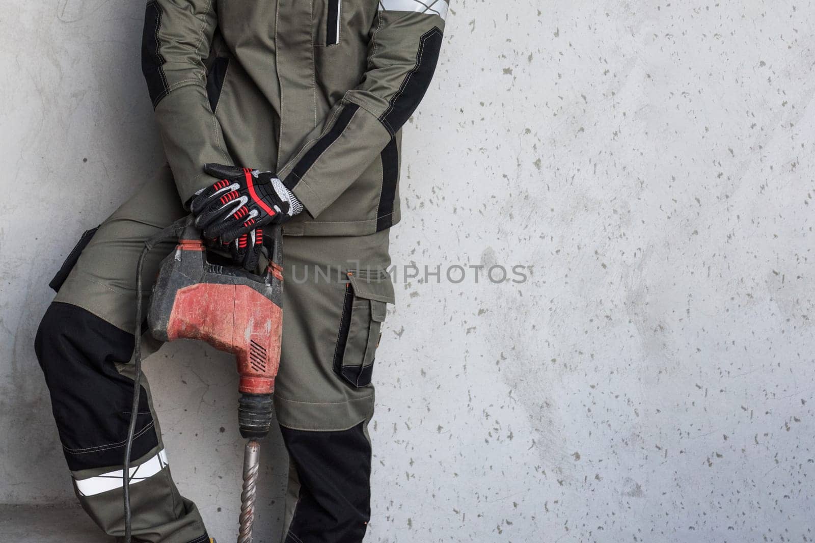 Man builder in workwear with electric hammer drill on grey concrete background with copyspace