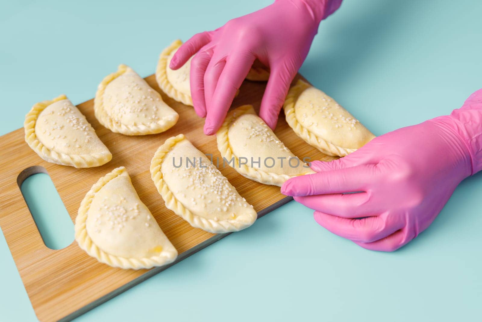 A baker's hands in a pink rubber gloves lays out raw samosa on the wooden board. Traditional Central Asian cuisine, frozen semi-finished products.