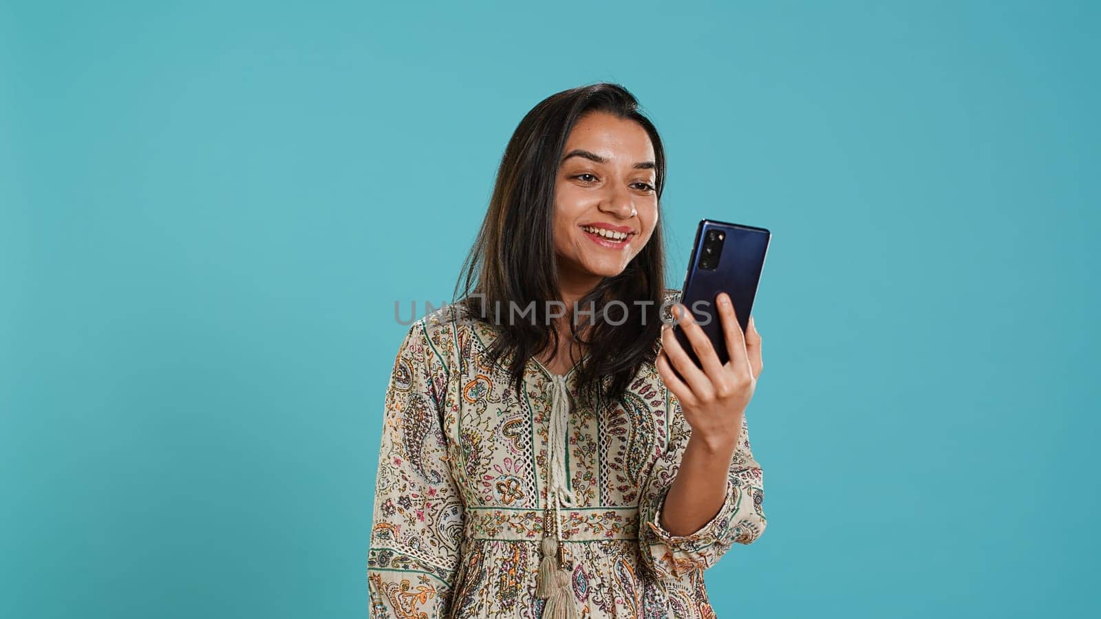 Smiling woman having friendly conversations during teleconference meeting using smartphone, studio background. Indian person having fun catching up with mates during online videocall, camera A