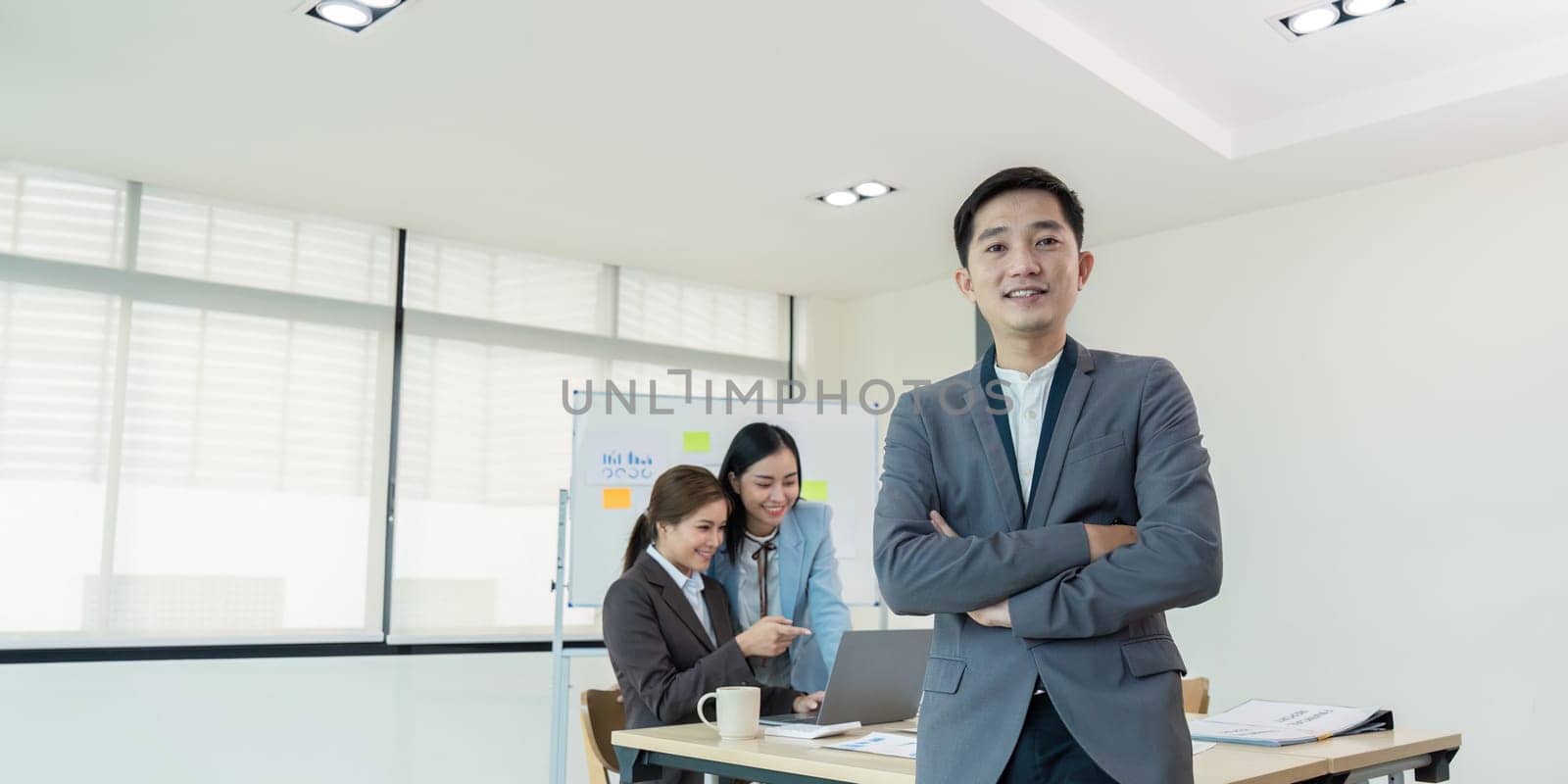 Confident business leader standing with arms crossed while his team collaborates on a laptop in the office, highlighting leadership and teamwork