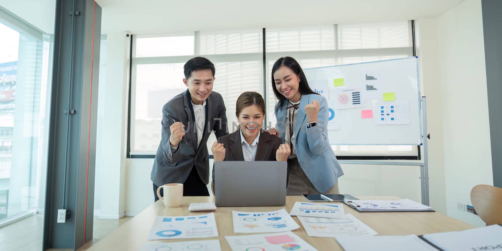 Three business colleagues in formal attire enthusiastically celebrating success while looking at a laptop in an office, emphasizing teamwork and achievement