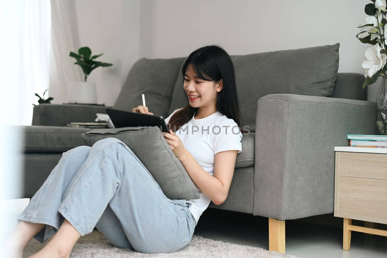 Cheerful female freelancer in casual clothes sitting on floor in living room and using digital tablet.
