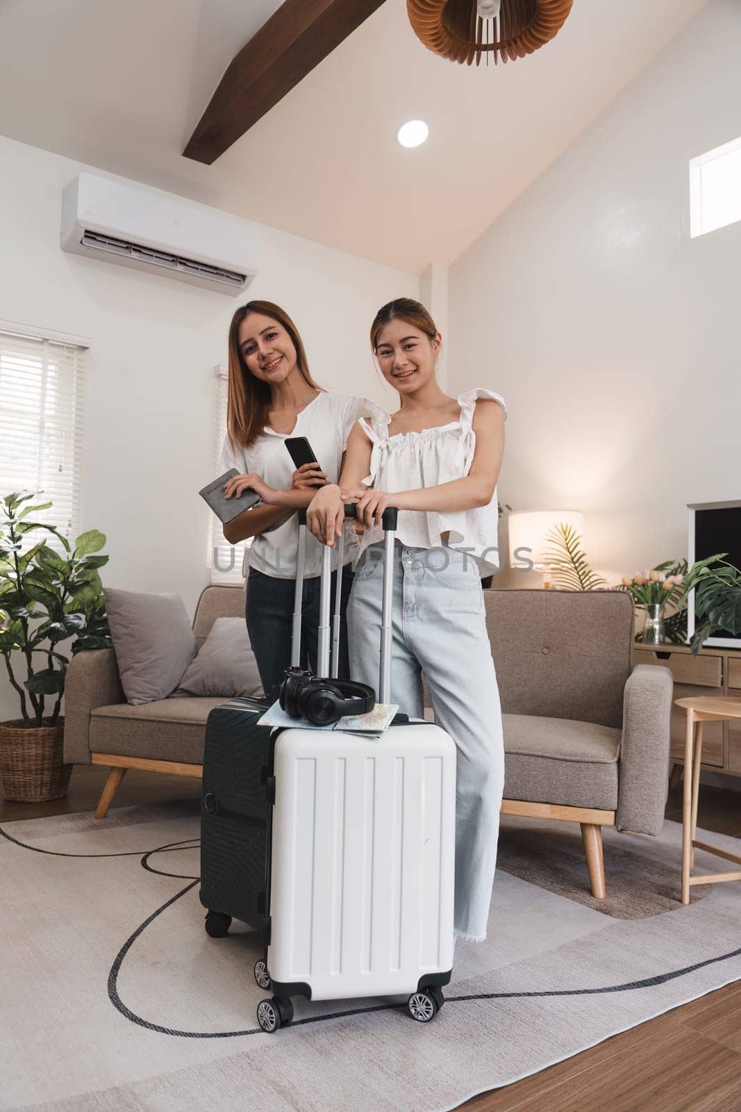 Two young women standing with suitcases and travel essentials in a living room, smiling and ready for a summer trip
