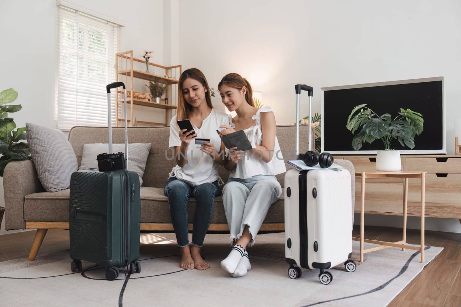 Two young women seated on a sofa, using a smartphone and credit card to book a hotel online, with suitcases nearby