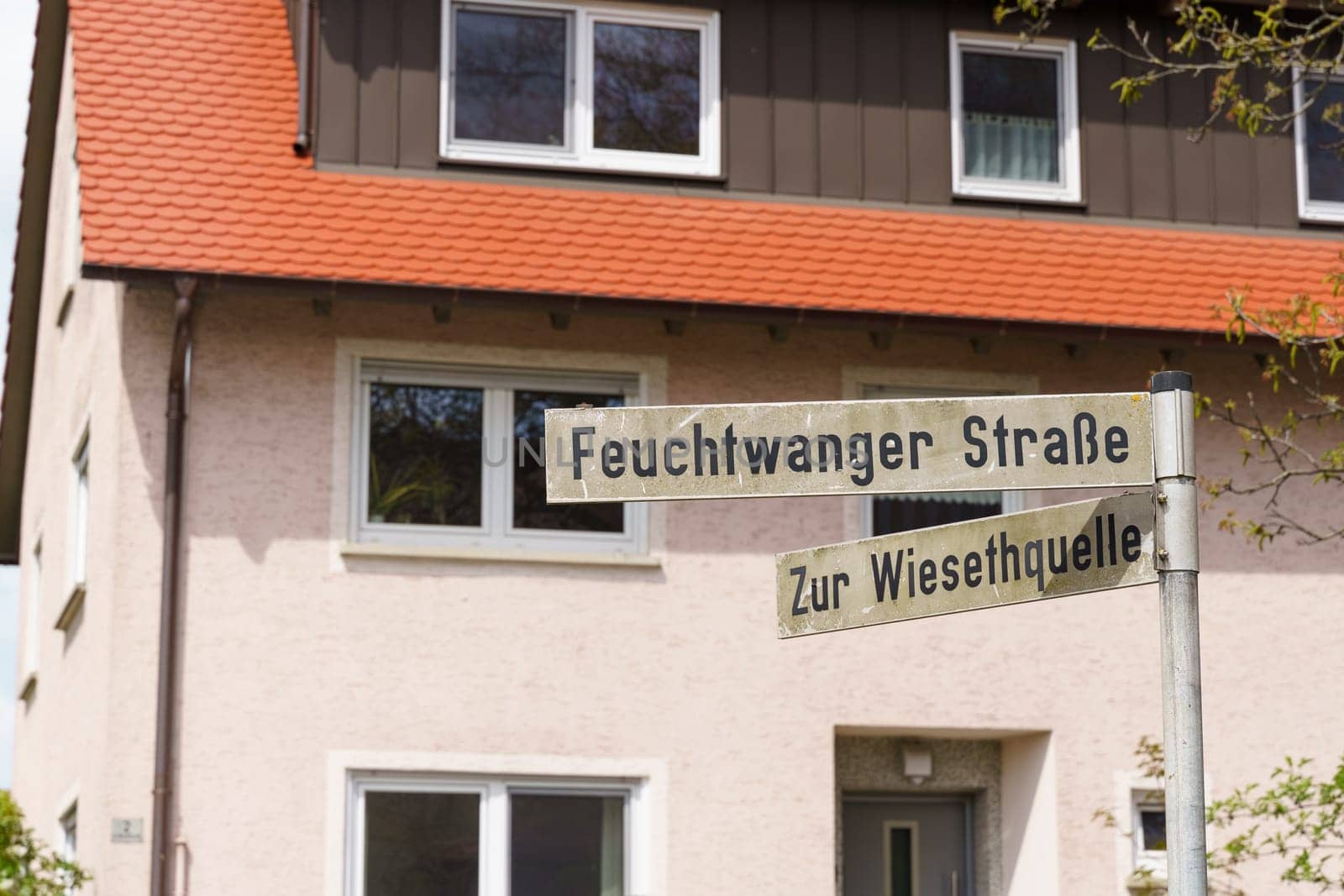 Two street signs point to Feuchtwanger Strabe and Zur Wiesehtquelle, in front of a white house with an orange roof.