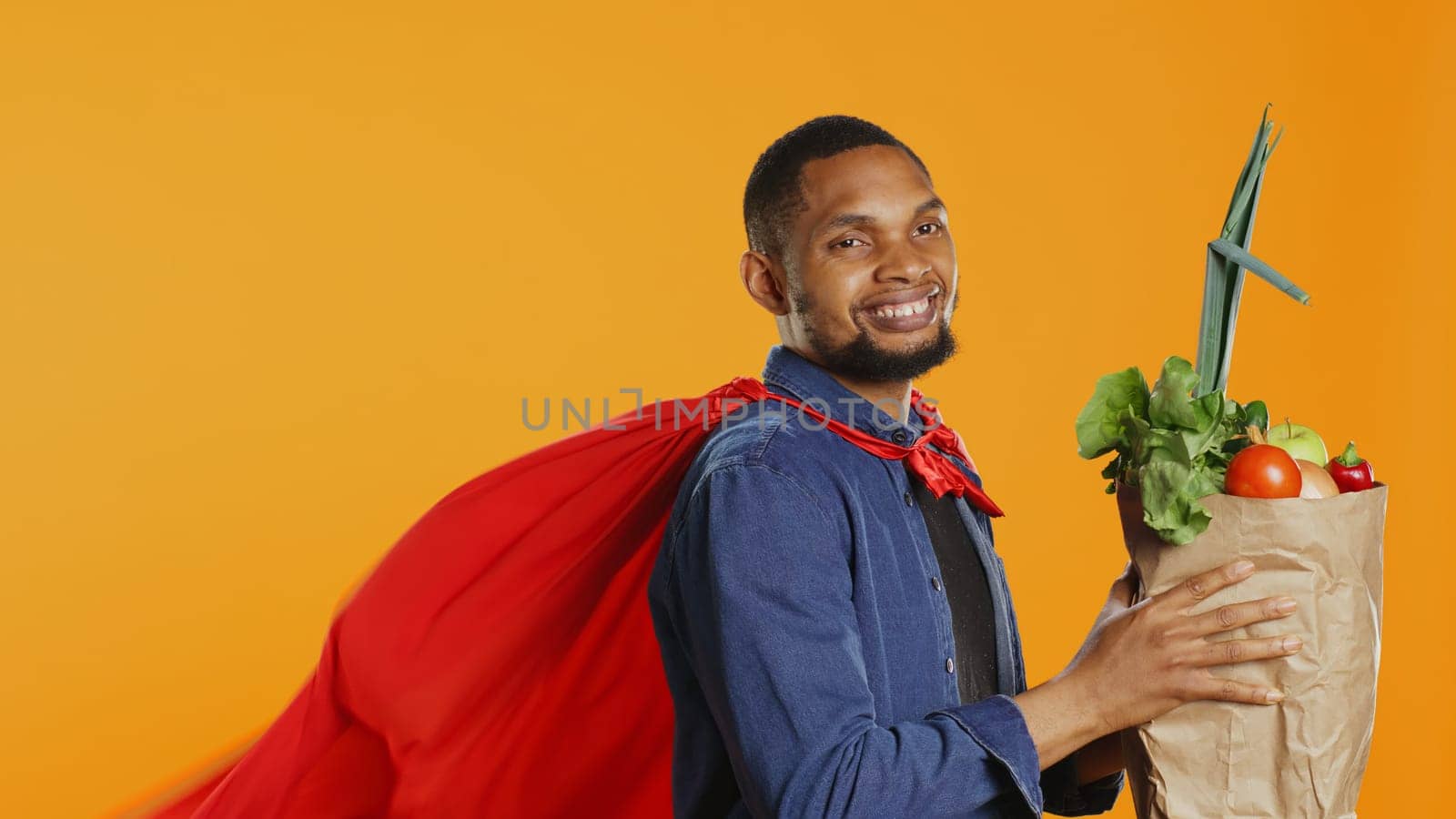 Young adult acting as a superhero with a red cape presenting homegrown produce in a paper bag, advocating for sustainable lifestyle concept. Person supporting organic food. Camera B.