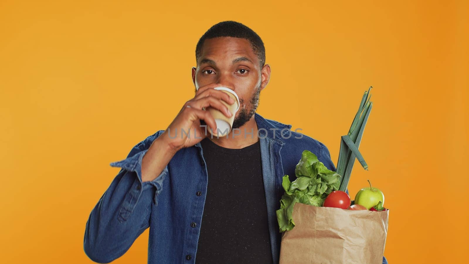 Relaxed carefree person drinking a coffee cup and carrying his groceries, going shopping at a local farmers market. Male model enjoying refreshment against orange background. Camera A.