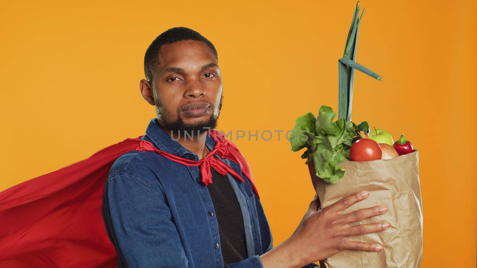 Young adult acting as a superhero with a red cape presenting homegrown produce in a paper bag, advocating for sustainable lifestyle concept. Person supporting organic food. Camera A.